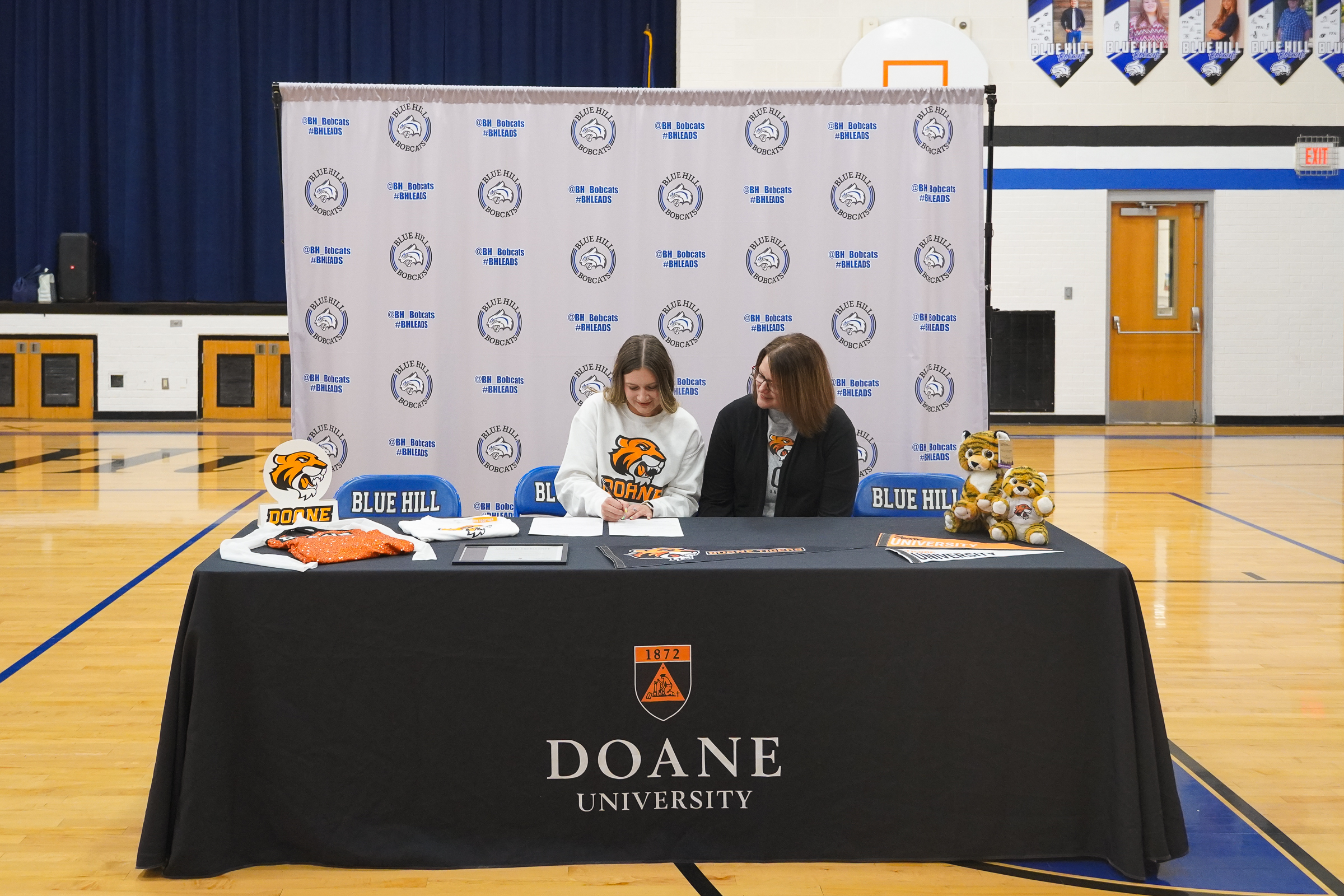 Two women sit behind a table draped with a black cloth that reads "Doane University." The woman on the left wears a Doane sweatshirt and is signing a document. Around them is Doane swag, including two stuffed tigers, several pennants, a shirt and a uniform for the Doane dance team.