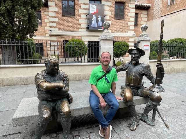 Josh Pope, wearing a green polo and jeans, sits between bronze statues of Sancho Panza and Don Quixote while traveling abroad in Madrid, Spain. Behind him is a brick building surrounded by a wrought-iron fence.