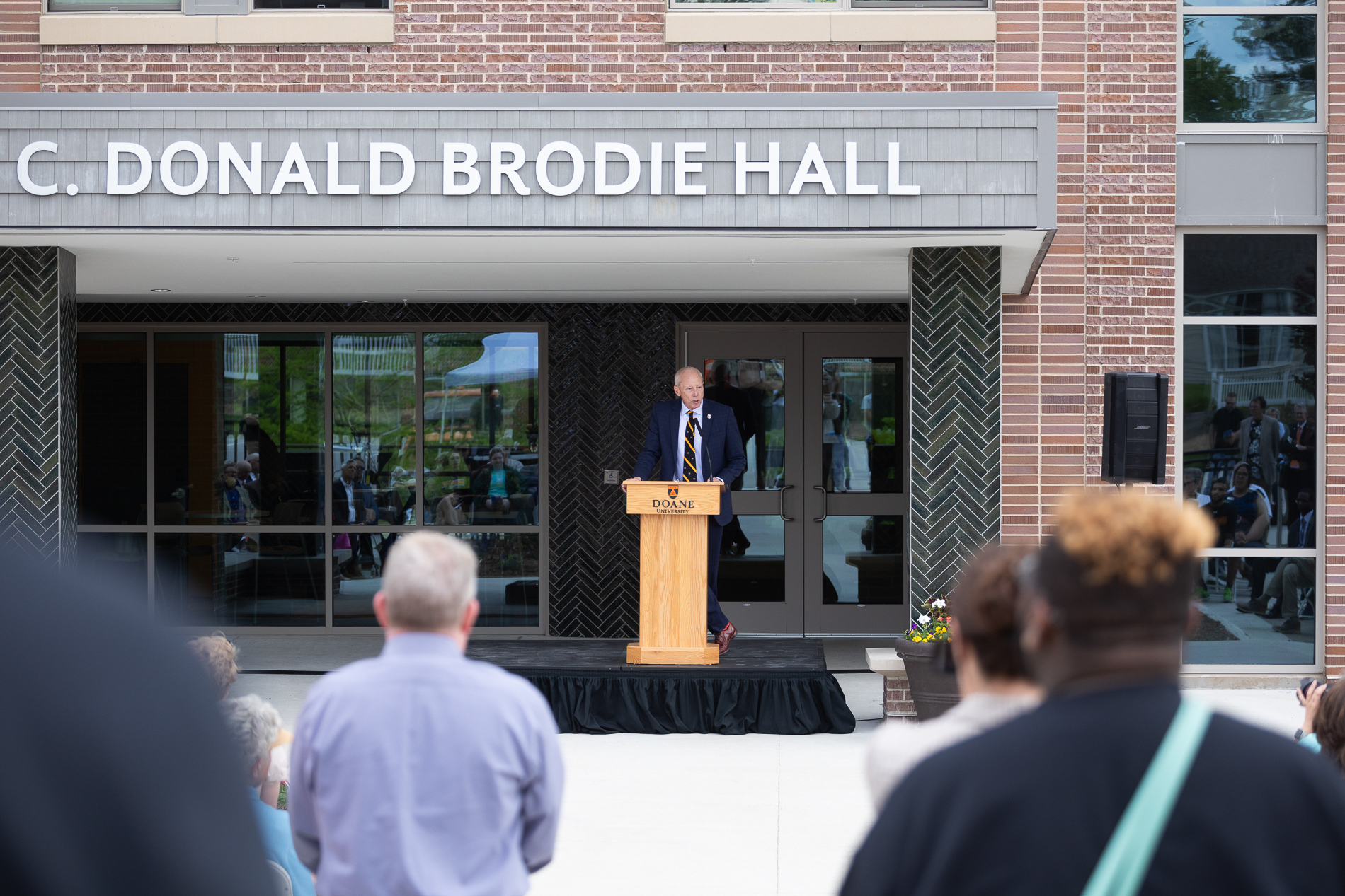 A man in a suit stands behind a wooden podium in front of an audience. Behind him is a building with a name reading "C. Donald Brodie Hall."