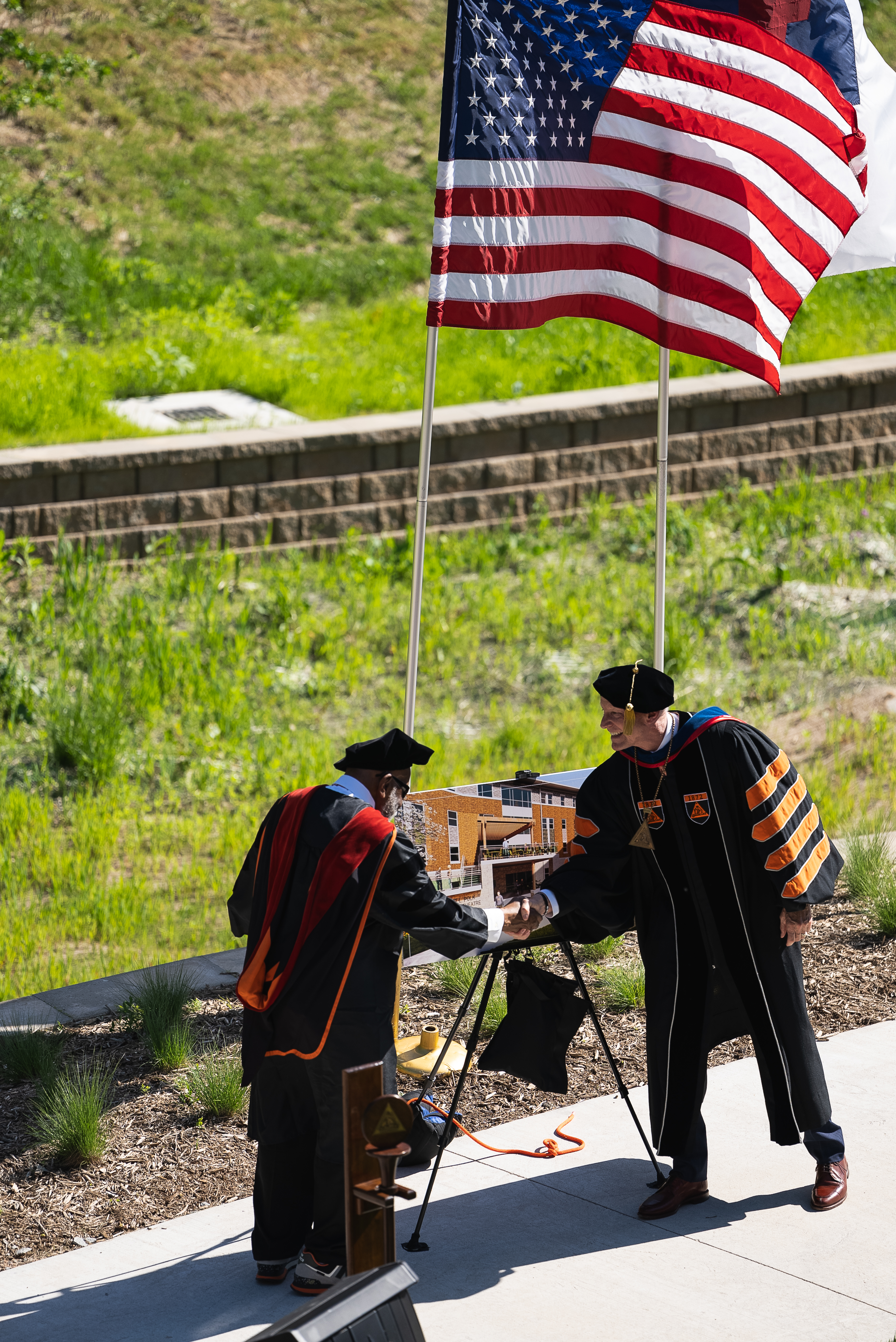 Two men in black academic robes shake hands in front of a poster showing a building. Behind them flies an American flag, with green plant growth in the background split by a brick retaining wall.