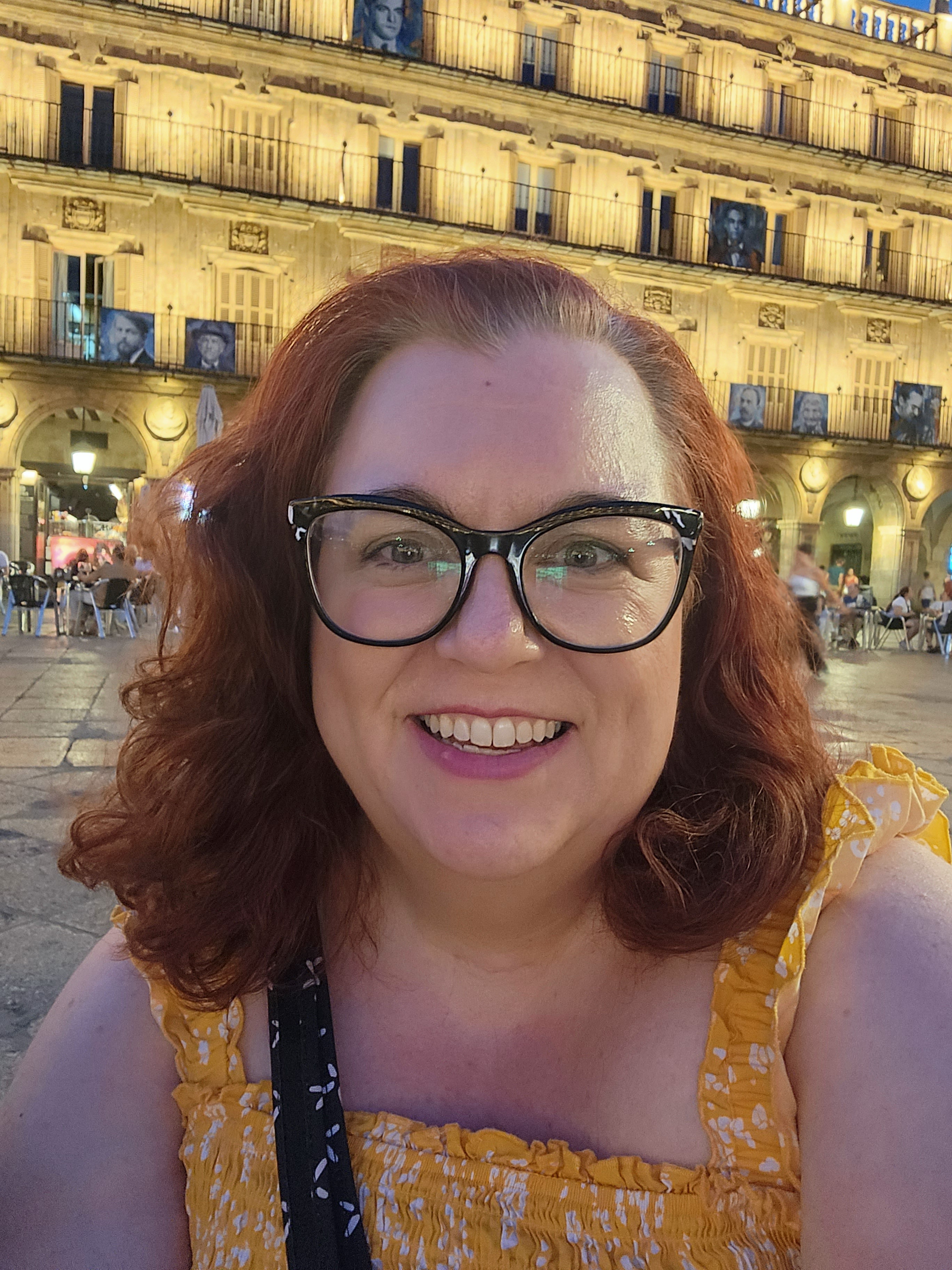 Melanie D'Amico takes a self-portrait while traveling abroad in Spain. She has curling, red hair and large, cat-eye glasses, and wears a yellow top.