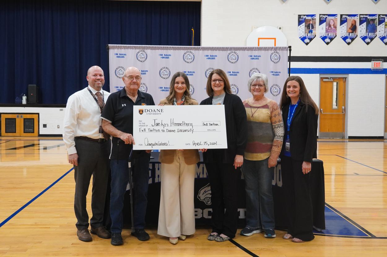 Six people stand in front of a white backdrop inside a school gymnasium. They're holding a giant check that reads "Pay to the order of Jaelyn Himmelberg; Full tuition to Doane University; Congratulations!"