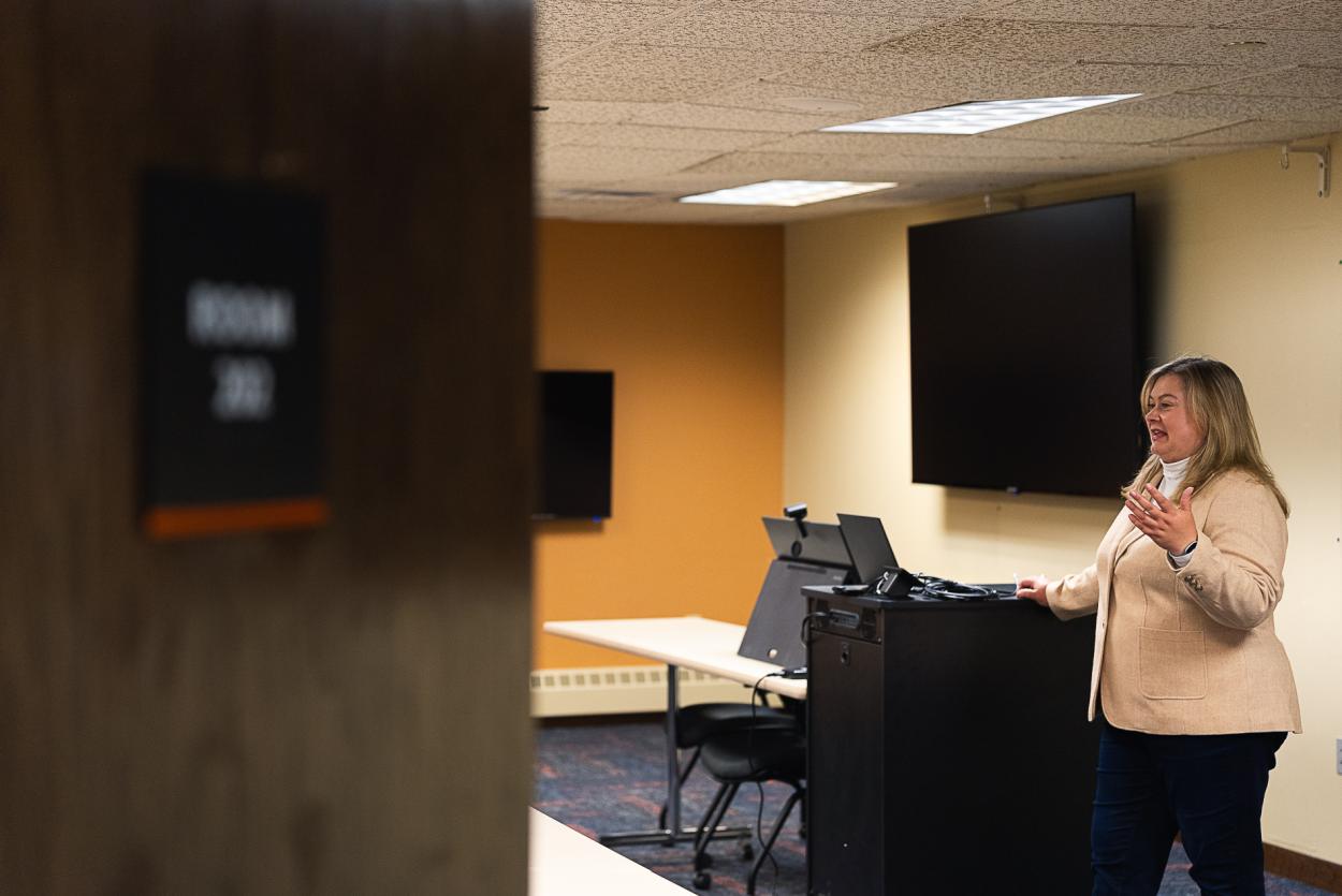 Dr. Jennifer Bossard speaks at the front of the College of Business's technology classroom, located in the Fred Brown Center in Lincoln. A partially closed door covers part of the image, while behind Bossard are two large televisions, a tablet and a cabinet with additional technology.