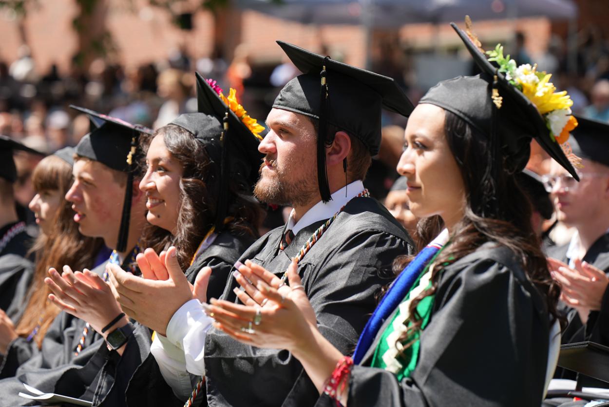 A row of graduates in black caps and gowns clap their hands, with rows of other students and families visible behind them.