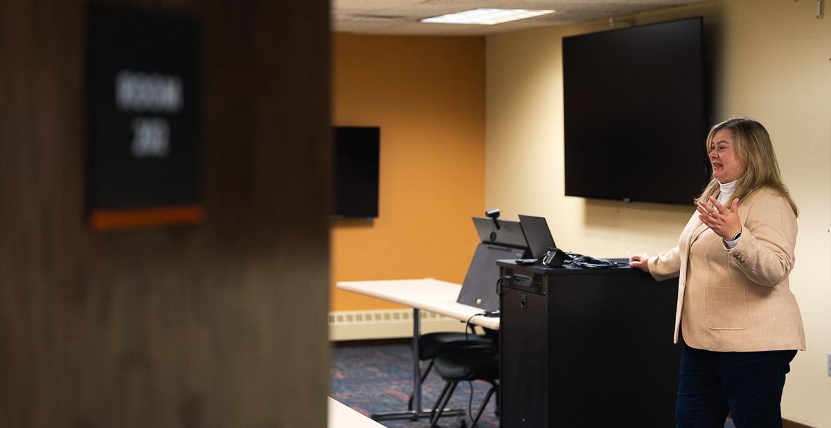 Dr. Jen Bossard speaks at the front of the College of Business's technology classroom, located in the Fred Brown Center in Lincoln. An open door partially covers the image, and behind Bossard are two large television screens, a tablet and a tech cabinet.